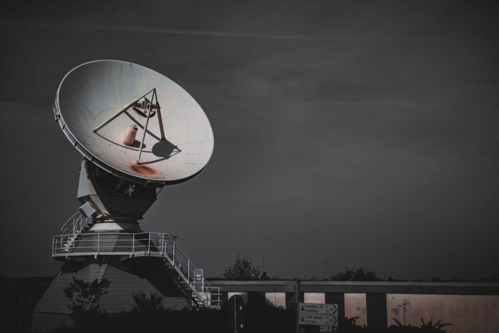 An imposing satellite dish is photographed against a dusky sky, featuring a prominent steel framework and the dish's parabolic shape designed for signal reception and transmission. The scene is composed with the satellite dish dominating the upper half of the frame, underscored by a building with directional signage partially visible below.