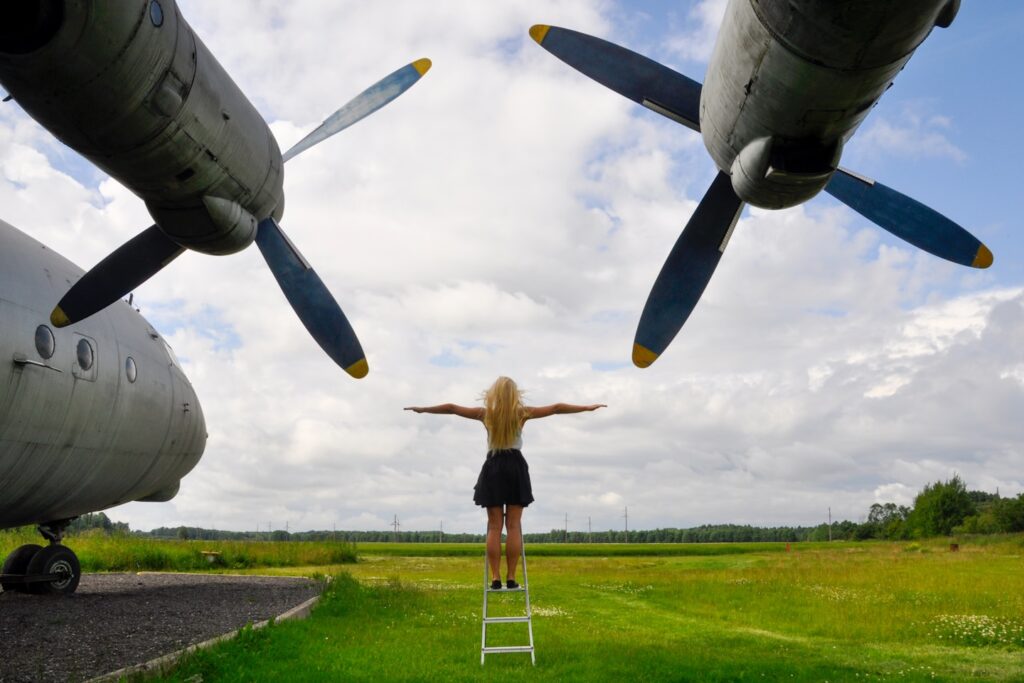 A blonde woman standing with outstretched arms beneath the massive propellers of a Tu-95 strategic bomber in Keila, Estonia, symbolizing freedom and the vast scale of aviation history."
