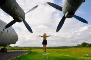 A blonde woman standing with outstretched arms beneath the massive propellers of a Tu-95 strategic bomber in Keila, Estonia, symbolizing freedom and the vast scale of aviation history."