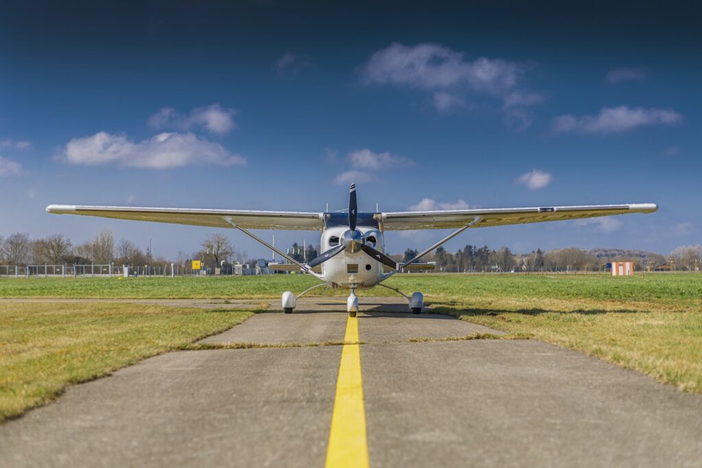 Ein leichtes Sportflugzeug vom Typ Cessna steht auf einer Start- und Landebahn. Die Aufnahme aus größerer Entfernung zeigt die Maschine mit vollständig sichtbaren Tragflächen und Propeller, während im Hintergrund ein kleines Flughafengebäude, grüne Wiesen und ein strahlend blauer Himmel im Sonnenlicht zu sehen sind. Photographer: Mark Ulm