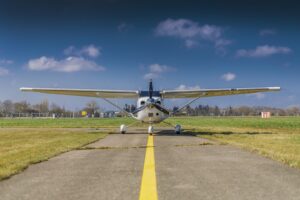 Ein leichtes Sportflugzeug vom Typ Cessna steht auf einer Start- und Landebahn. Die Aufnahme aus größerer Entfernung zeigt die Maschine mit vollständig sichtbaren Tragflächen und Propeller, während im Hintergrund ein kleines Flughafengebäude, grüne Wiesen und ein strahlend blauer Himmel im Sonnenlicht zu sehen sind. Photographer: Mark Ulm